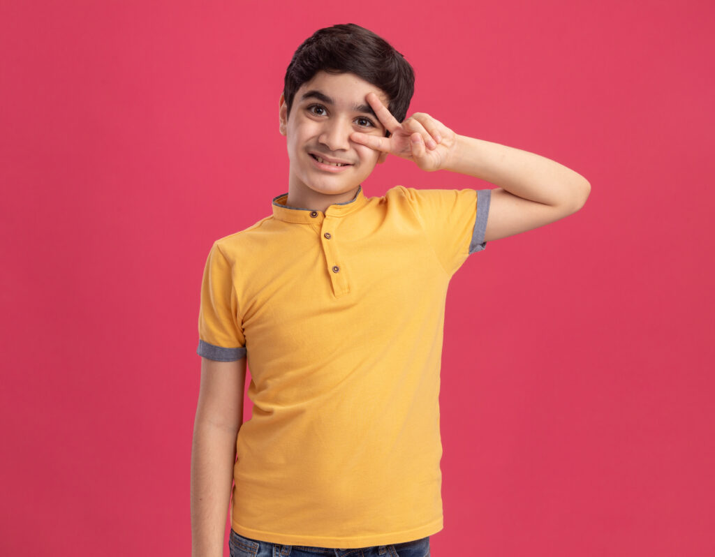 smiling young caucasian boy looking at camera showing v sign symbol near eye isolated on pink background with copy space smiling young caucasian boy looking at camera showing v sign symbol near eye isolated on pink background with copy space